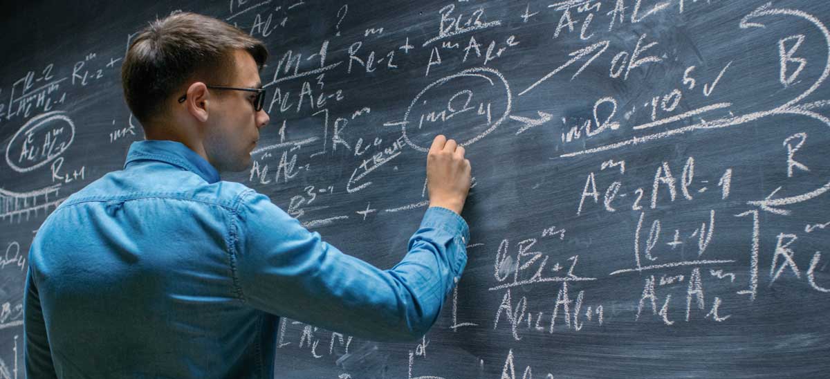 A man with glasses in a blue shirt writing physics equations on a chalkboard