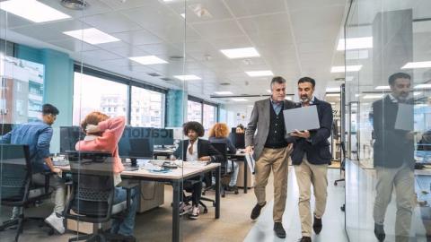 Two men walking through a busy office space, looking at a laptop