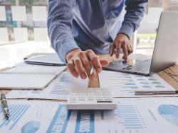 A man using a calculator that is sitting on a table along with many papers covered in charts and graphs